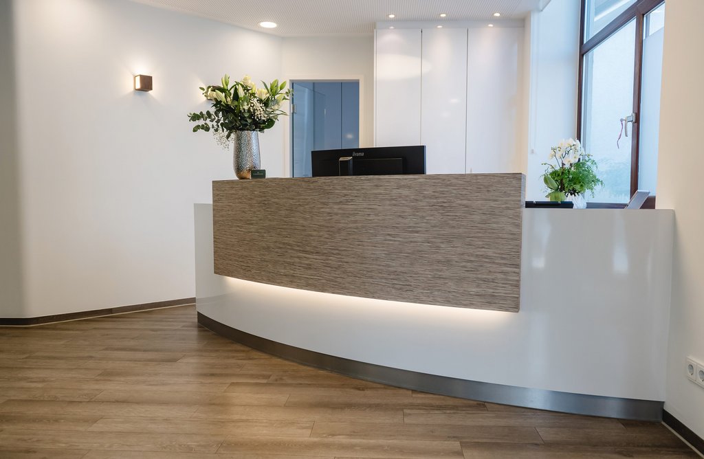 Moderne Empfangstheke in der zahnmedizinischen Praxis Dr. Sigmund & Kollegen mit Holzverkleidung, Rundung und Blumendekoration, beleuchtet durch indirektes Licht und natürliches Tageslicht.