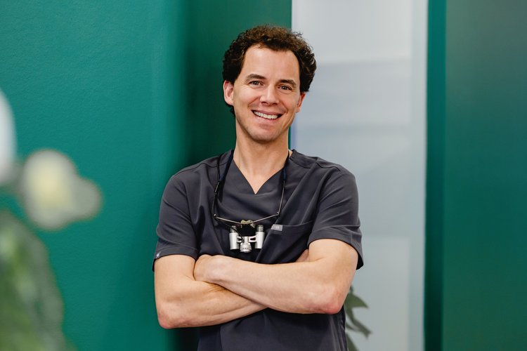 Dr. med. dent. Fabian Sigmund steht lächelnd mit verschränkten Armen in dunkelgrauer Berufskleidung und eine Zahnlupe um den Hals vor neutralem Hintergrund.