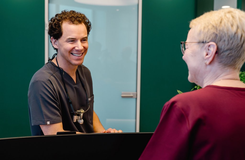 Dr. Fabian Sigmund berät lächelnd eine ältere Patientin am Empfang der zahnmedizinischen Praxis in Inning.