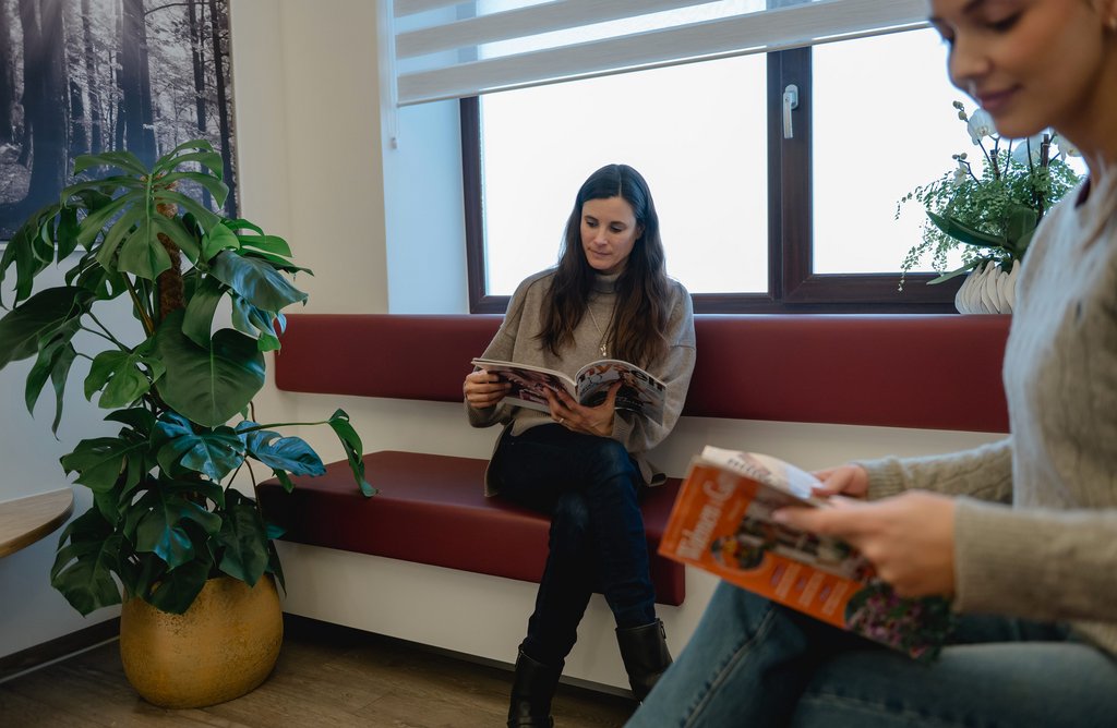 Zwei Frauen sitzen im Wartezimmer der Zahnarztpraxis Dr. Sigmund & Kollegen und lesen Zeitschriften, im Raum steht eine große grüne Pflanze.