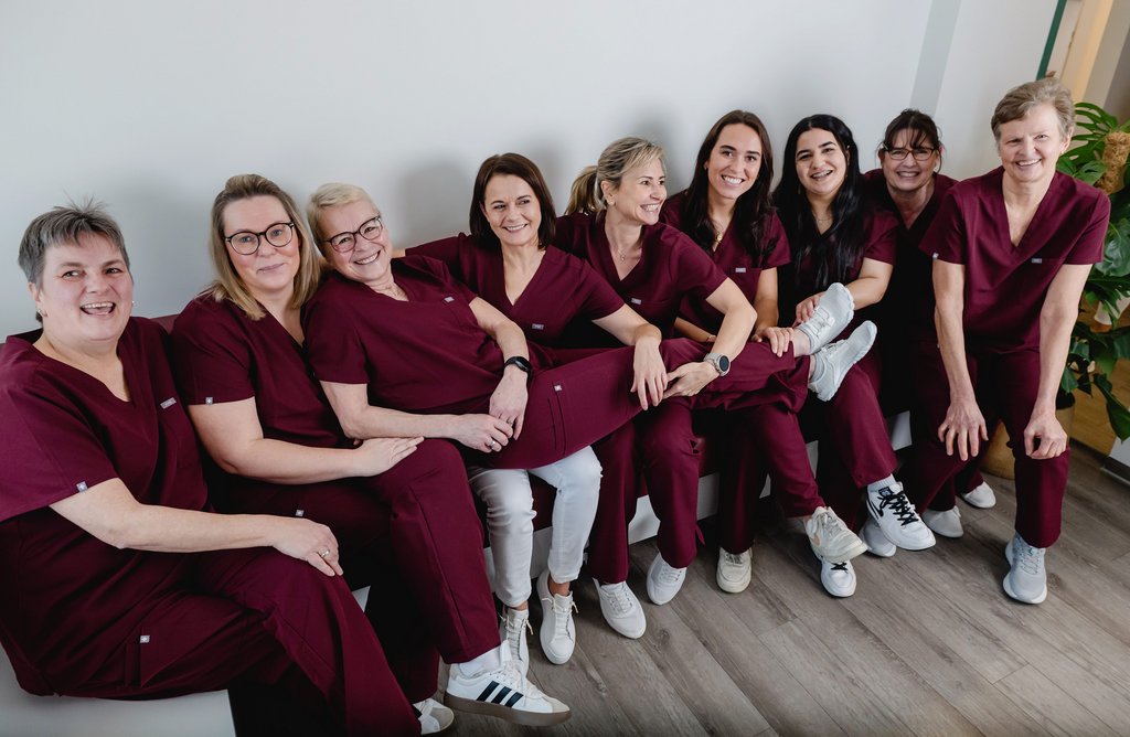 Das Praxisteam der Zahnarztpraxis Dr. Sigmund & Kollegen in einheitlicher, dunkelroter Arbeitskleidung sitzt lachend nebeneinander, eine Frau wird von den anderen gehalten.
