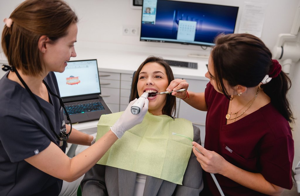 Eine Patientin sitzt im Behandlungsstuhl, während Maja Schreiner und eine ZFA ihre Zähne untersuchen und mit zahnmedizinischen Instrumenten arbeiten.