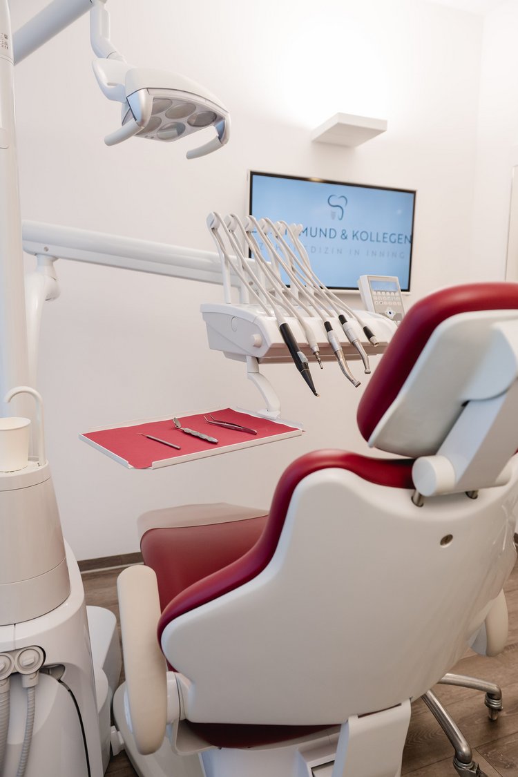 Behandlungszimmer in der zahnmedizinischen Praxis Dr. Sigmund & Kollegen mit einem roten Zahnarztstuhl, verschiedenen zahnmedizinischen Instrumenten und einem Bildschirm an der Wand.