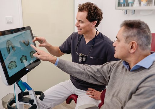Dr. Fabian Sigmund erklärt einem Patienten anhand von 3D-Bildern auf einem Bildschirm die Schritte der Gesamtsanierung des Gebisses.