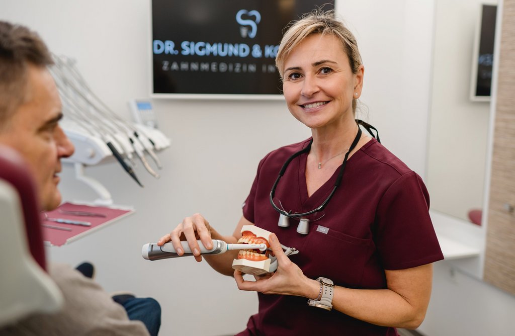 Eine ZFA der Praxis Dr. Sigmund & Kollegen erklärt einem Patienten mit einem Zahnmodell und einer elektrischen Zahnbürste die Zahnpflege.