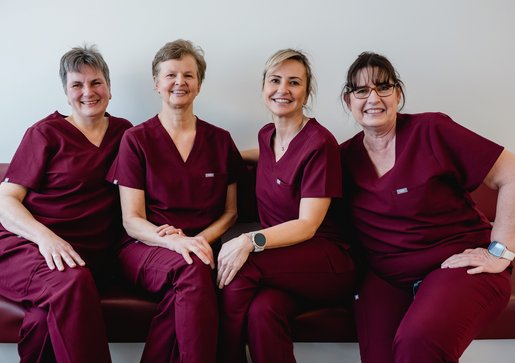 Vier weibliche ZFA der zahnmedizinischen Praxis Dr. Sigmund & Kollegen in weinroten medizinischen Kitteln sitzen nebeneinander auf einer Bank und lächeln freundlich in die Kamera.