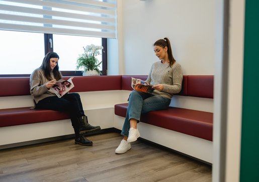 Zwei junge Frauen sitzen auf einer roten Eckbank und lesen jeweils ein Magazin im Wartezimmer der Praxis Dr. Sigmund & Kollegen.